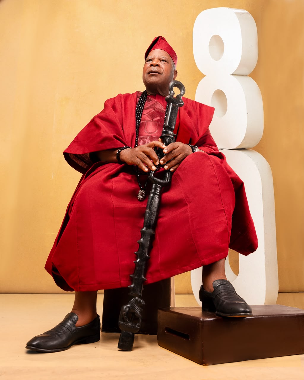 Elder in a red agbada with a staff beside an 80th birthday sign